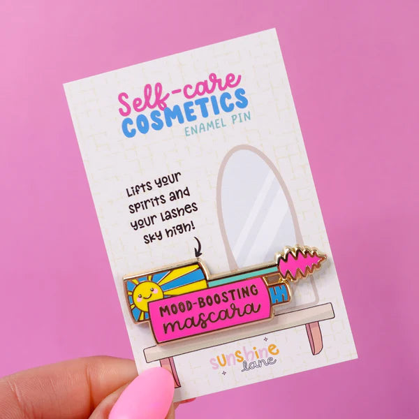 A hand holds a self-care gift enamel pin on a card that reads Mood-Boosting Mascara, featuring colourful illustrations set against a pink background.