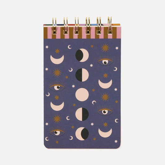 A spiral-bound celestial notebook with a purple cover, embellished with moon phases, stars, and eye illustrations in gold, black, and white.