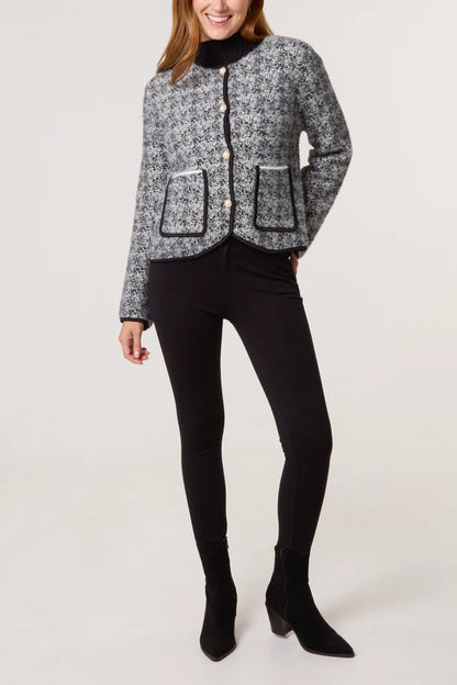 A person wearing a textured bouclé knit cardigan in grey and black with front pockets, paired with black trousers and black ankle boots, stands against a plain background.