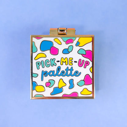 A square make-up palette with a gold border and colourful abstract shapes on the lid, labelled PICK-ME-UP palette, ideal as a self-care gift, set against a light blue background.