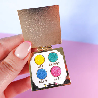 A hand holds an open, gold-coloured eyeshadow palette pin featuring four glittery circles labelled Joy (yellow), Energy (teal), Calm (blue), and Hope (pink)—a delightful self-care gift.