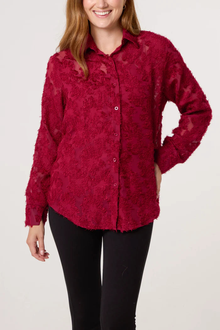 A woman wears a textured, long-sleeved red mesh shirt with a collar and button-up front, paired with black trousers, standing against a plain light background.