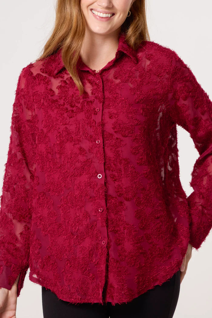 A woman wearing a textured, sheer dark red mesh shirt with a collar, standing against a plain backdrop.