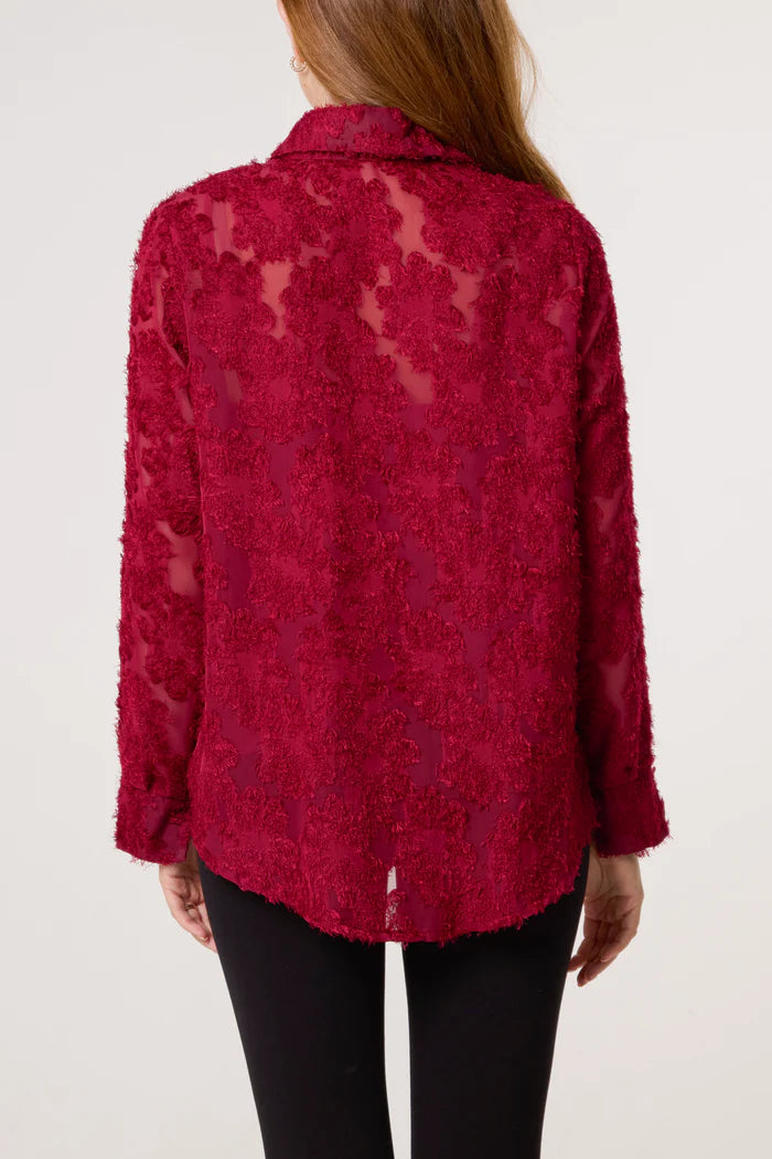 A person seen from behind wearing a long-sleeved, sheer red mesh top with floral patterns. The lightweight polyester blouse is teamed with black trousers.