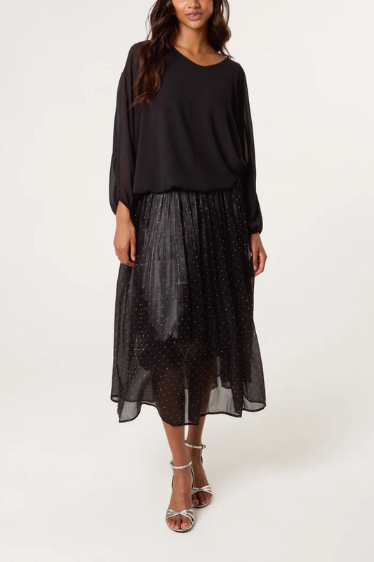 Woman wearing a loose black long-sleeved top and a sheer black shimmer midi skirt with small dots, paired with silver strappy heels, standing against a plain background.