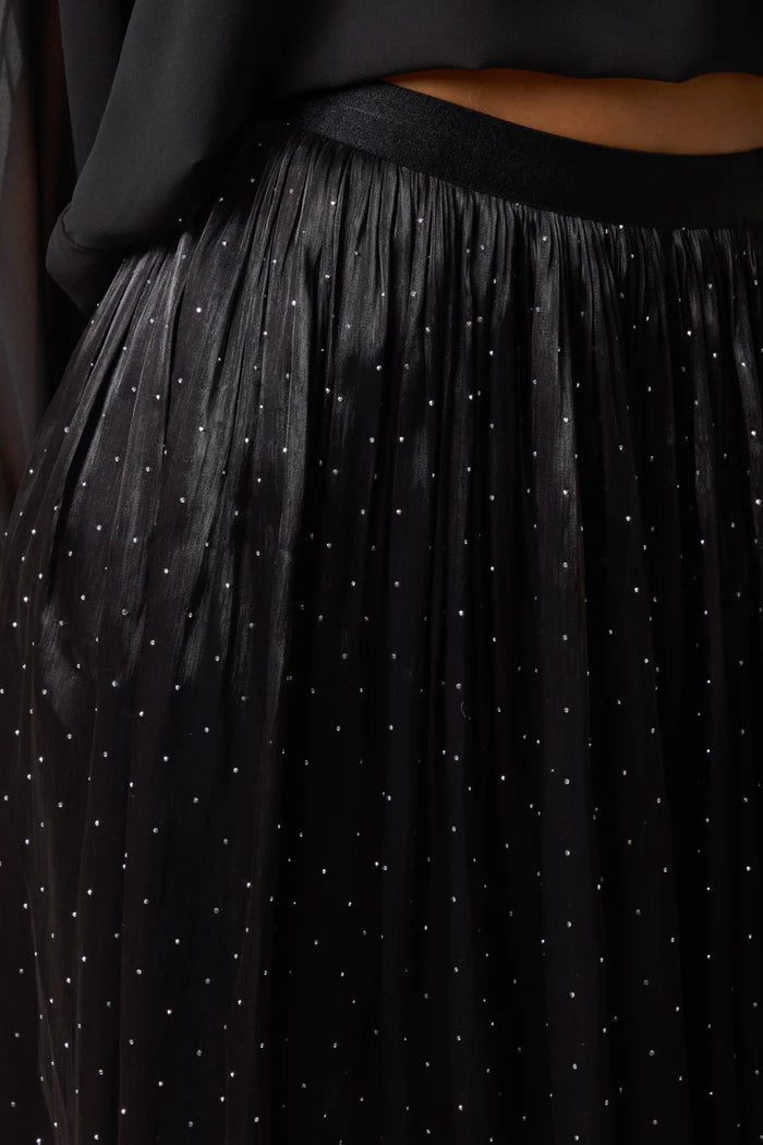 A close-up of a black shimmer midi skirt featuring sheer, glimmering fabric with small white polka dots, styled with a black top for an elegant evening ensemble.