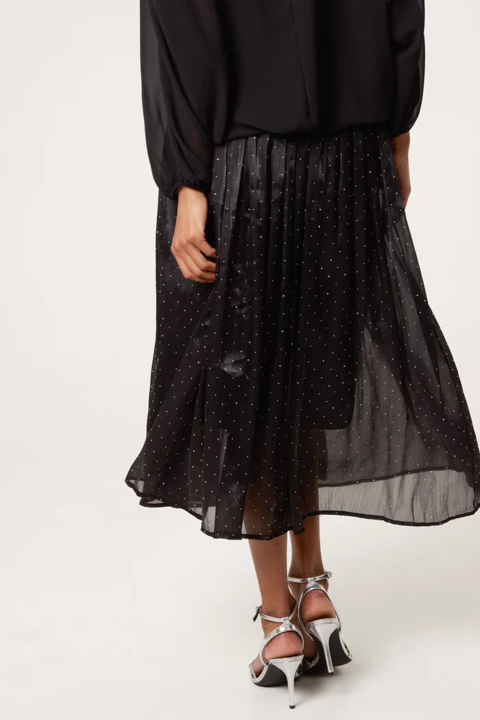 A person dressed in a black long-sleeved top, a shimmery midi skirt with small white dots, and silver high-heeled sandals, seen from behind.