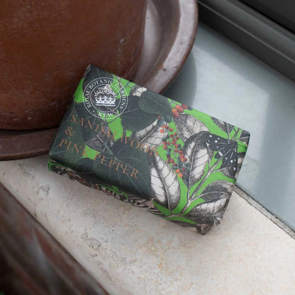 A vegan sandalwood soap, wrapped in green and black botanical paper labelled “Sandalwood & Pink Pepper”, sits beside a brown pot on the windowsill.