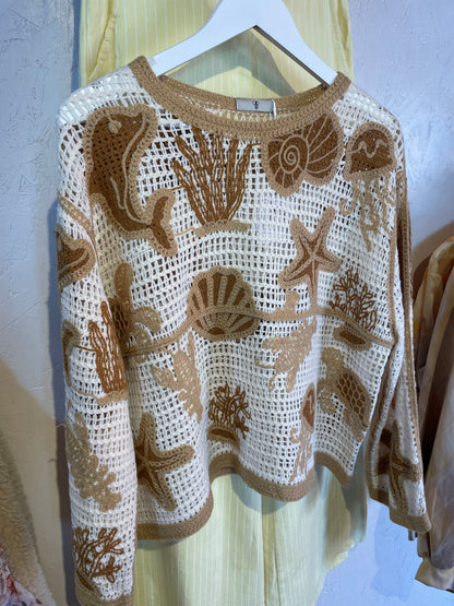 A cream sealife jumper featuring brown and beige sea-inspired motifs—shells, fish, coral, and starfish—hangs on a white hanger against a pale background.