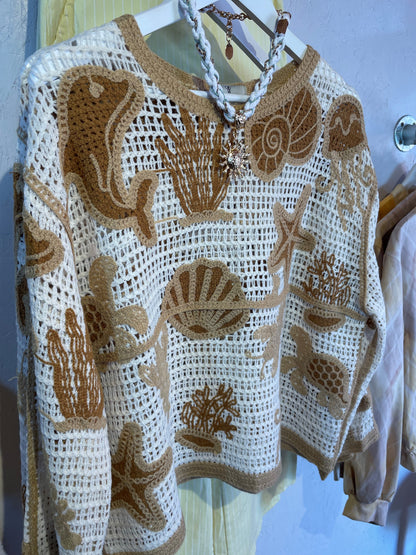 A cream sealife jumper crocheted with tan and white yarn displays ocean-themed patterns such as dolphins, shells, starfish, coral, and seaweed, evoking coastal charm as it hangs on a white hanger.