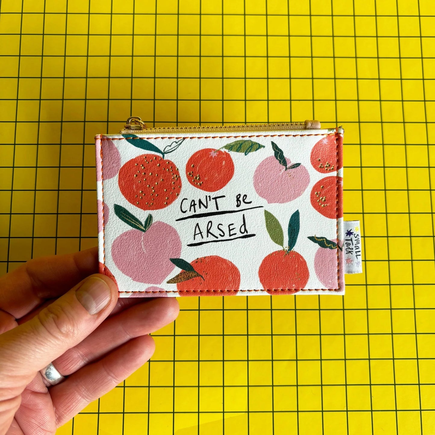 A hand holds a cheeky zipped purse featuring a fruit pattern and the words CANT BE ARSED on a yellow grid background—a perfect sarcastic present.