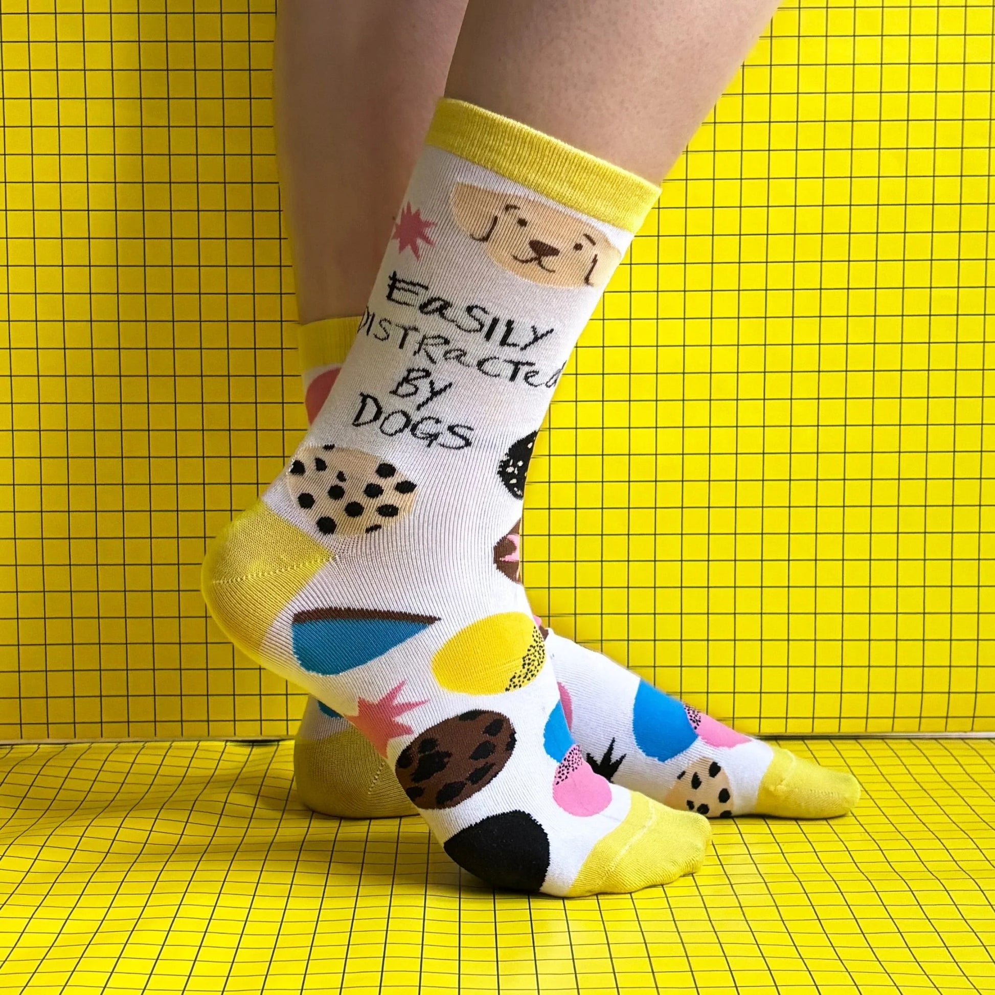 Individual wearing colourful dog lover socks featuring dog illustrations and the phrase "Easily distracted by dogs" set against a yellow, grid-patterned background.