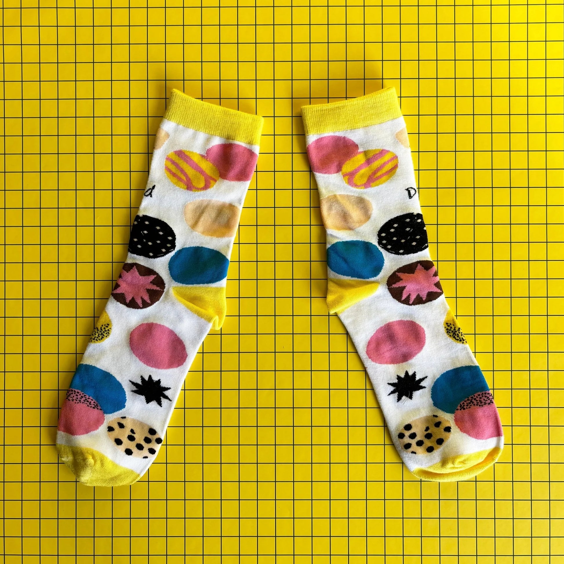 A pair of white bamboo socks with colourful circle patterns is shown on a yellow grid background.