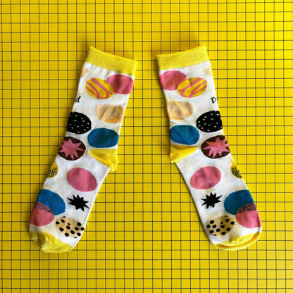 A pair of white bamboo socks with colourful circle patterns is shown on a yellow grid background.
