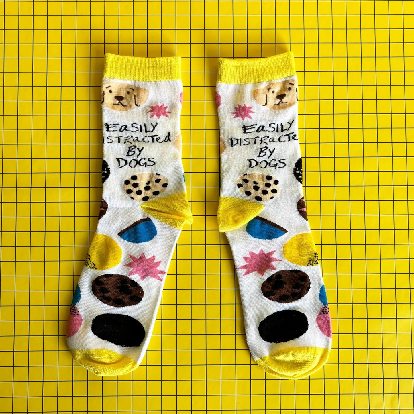 A pair of socks for dog lovers, featuring dog illustrations and the phrase "Easily Distracted By Dogs," displayed on a yellow grid background.