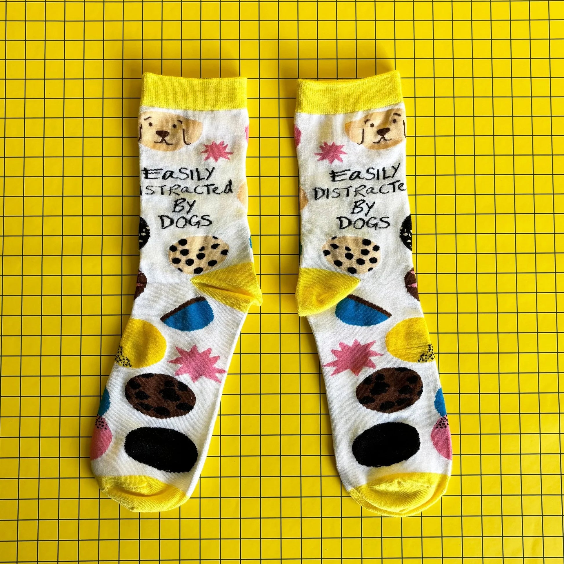 A pair of socks for dog lovers, featuring dog illustrations and the phrase "Easily Distracted By Dogs," displayed on a yellow grid background.
