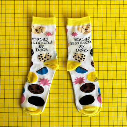 A pair of socks for dog lovers, featuring dog illustrations and the phrase "Easily Distracted By Dogs," displayed on a yellow grid background.