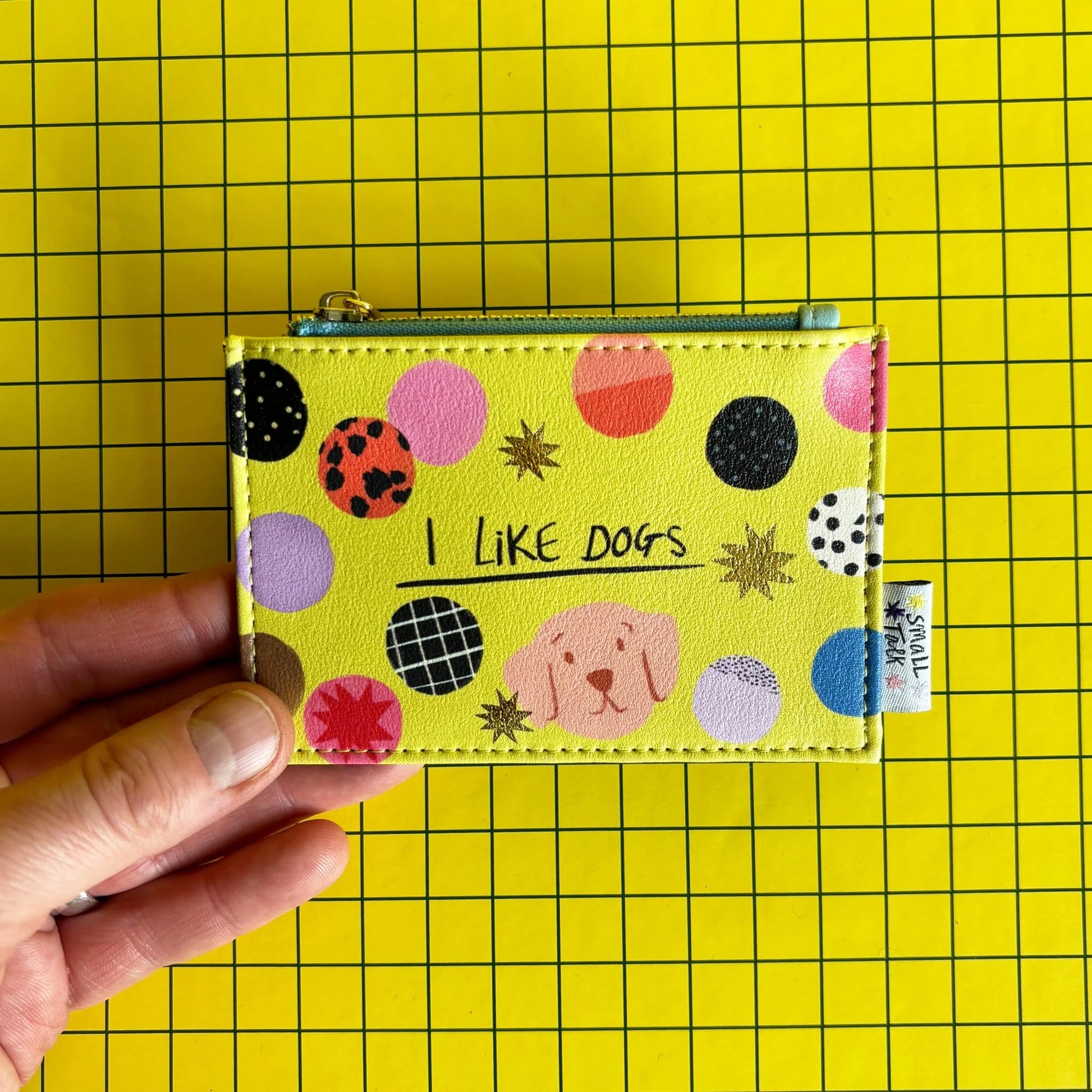 A hand holds a yellow zip coin purse featuring colourful circles, a small dog illustration, and the words I LIKE DOGS on a yellow grid background—ideal as a gift for dog lovers.