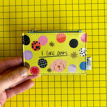 A hand holds a yellow zip coin purse featuring colourful circles, a small dog illustration, and the words I LIKE DOGS on a yellow grid background—ideal as a gift for dog lovers.