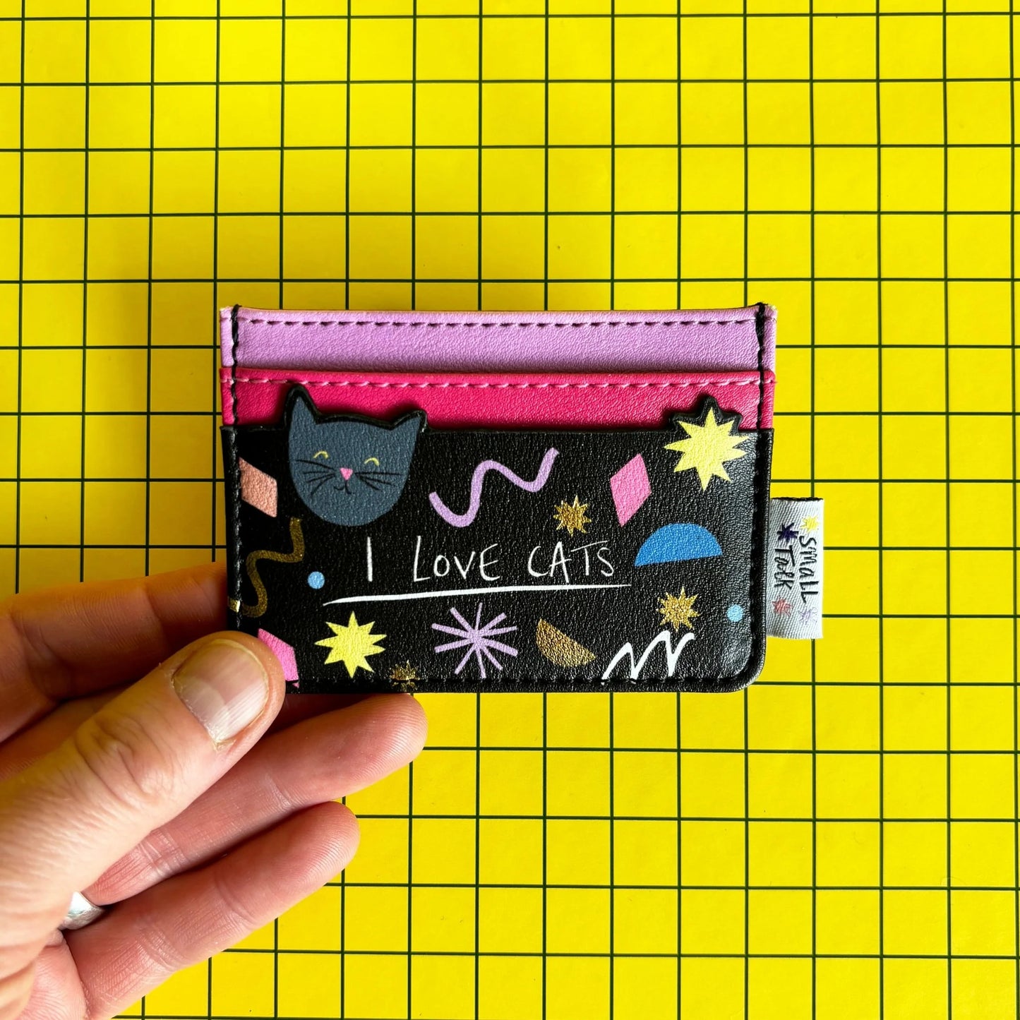 A hand holds a compact PVC cat card holder featuring cat illustrations, pink and red slots, and the words LOVE CATS, set against a yellow grid background.