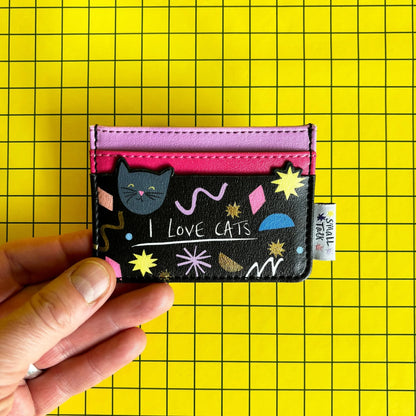 A hand holds a compact PVC cat card holder featuring cat illustrations, pink and red slots, and the words LOVE CATS, set against a yellow grid background.