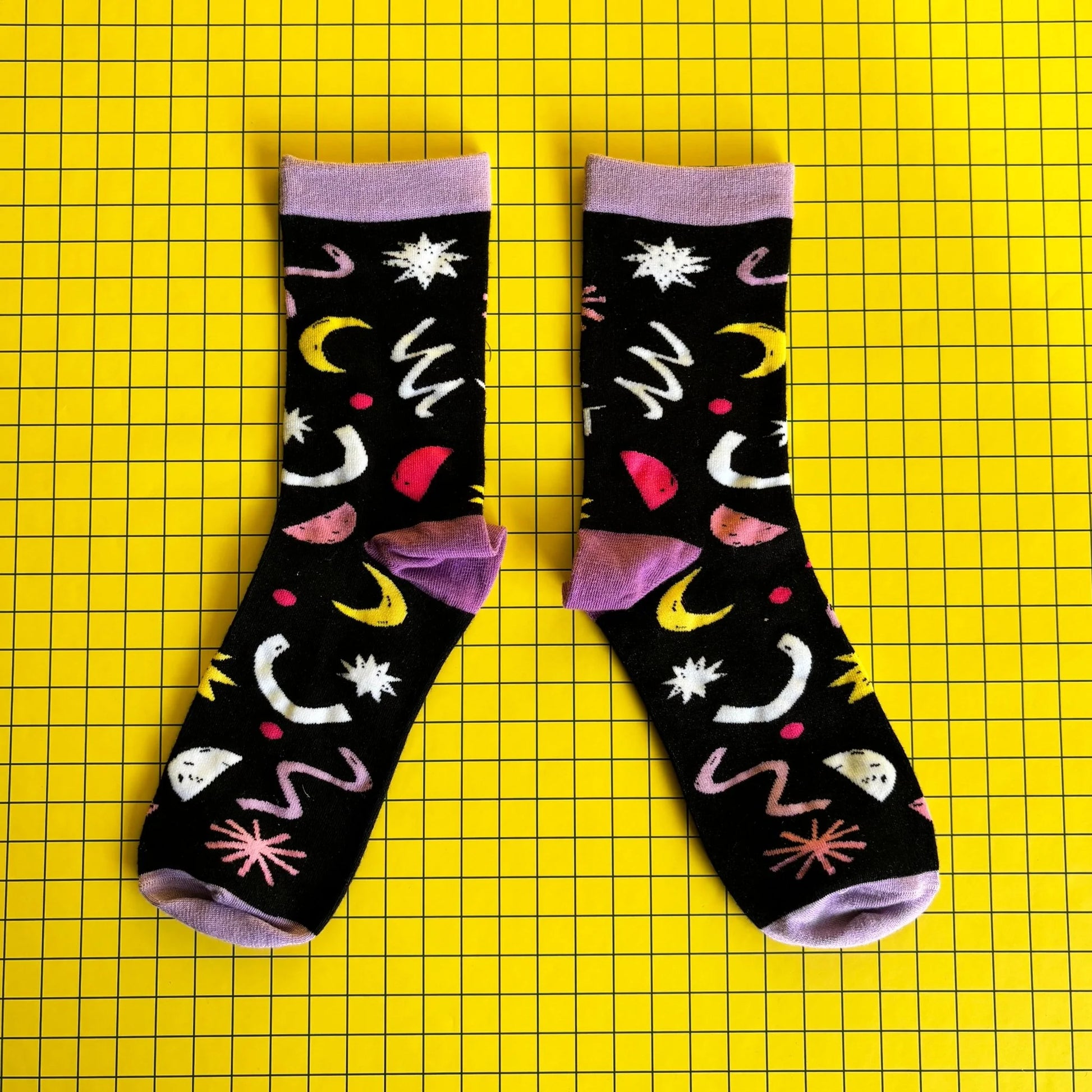 A pair of black bamboo socks featuring colourful abstract designs, including moons and stars, set against a yellow grid background—ideal as a quirky gift for cat lovers.