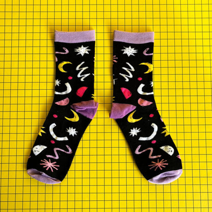 A pair of black bamboo socks featuring colourful abstract designs, including moons and stars, set against a yellow grid background—ideal as a quirky gift for cat lovers.