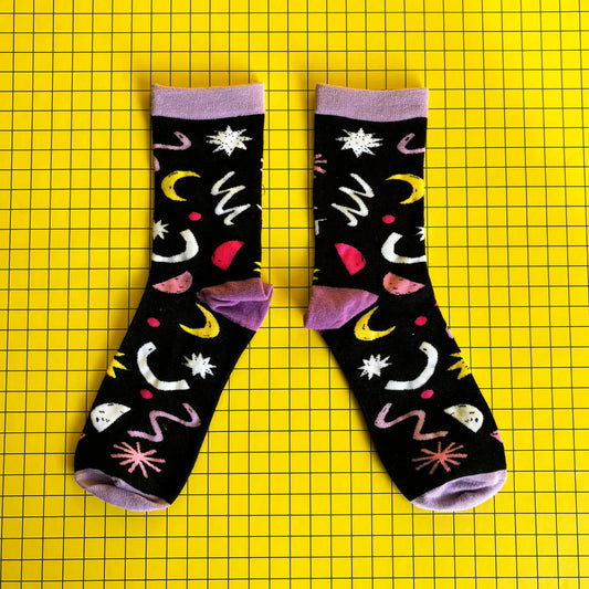 A pair of black bamboo socks featuring colourful abstract designs, including moons and stars, set against a yellow grid background—ideal as a quirky gift for cat lovers.