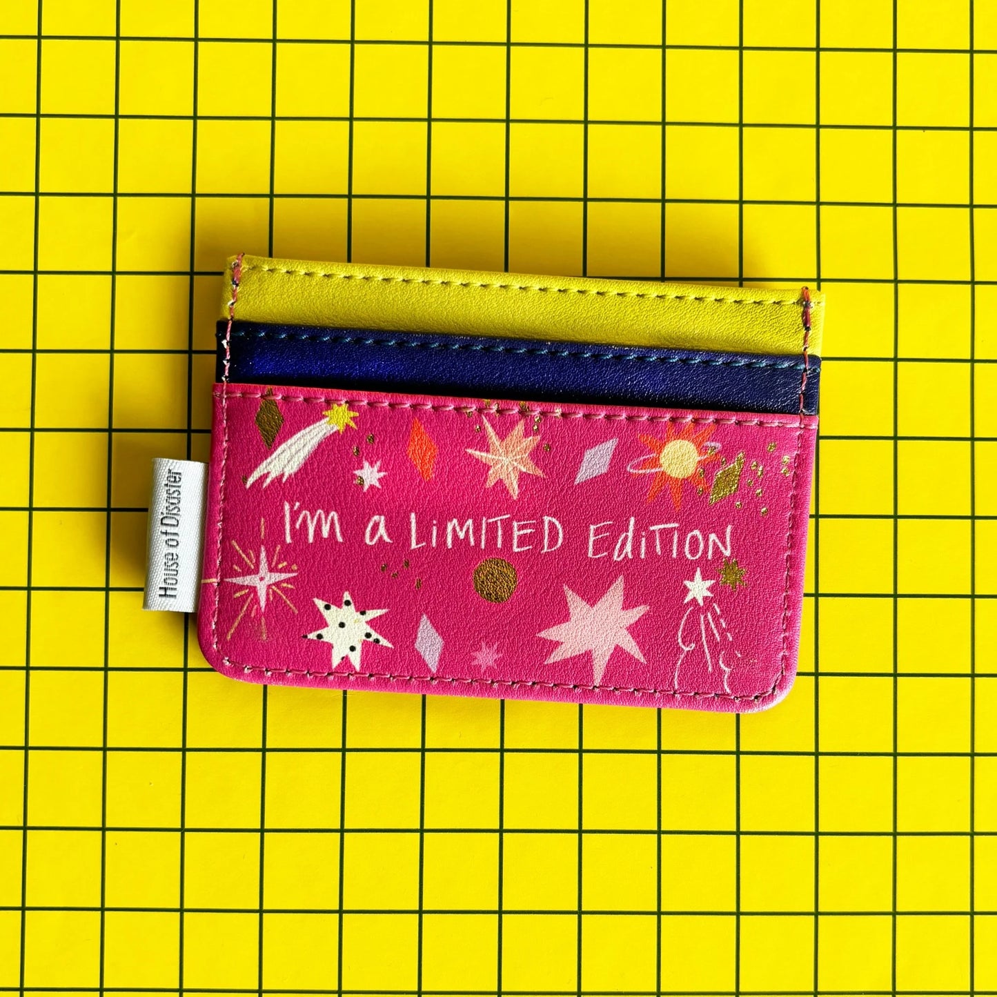 A quirky, limited edition cardholder featuring the text "I'm a LIMITED Edition" rests on a yellow grid background. The compact PVC holder is pink, decorated with stars and planets.