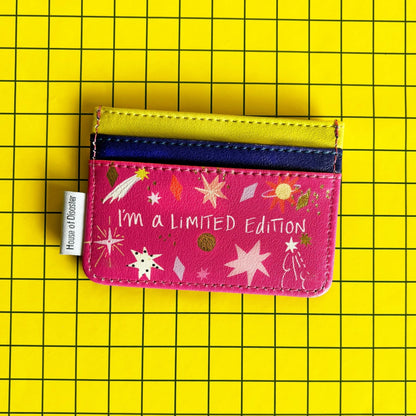 A quirky, limited edition cardholder featuring the text "I'm a LIMITED Edition" rests on a yellow grid background. The compact PVC holder is pink, decorated with stars and planets.