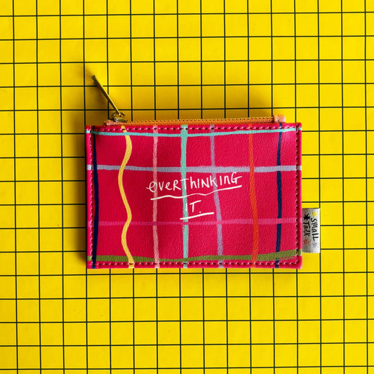 A pink tartan pocket-sized purse with OVERTHINKING IT written on the front, set against a yellow grid background—this overthinking purse makes a clever gift for friends who appreciate quirky accessories.