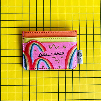 A pink, pocket-sized card holder featuring colourful rainbow designs and the word OVERWHELMED, set against a yellow grid background—ideal as a quirky gift for mates.