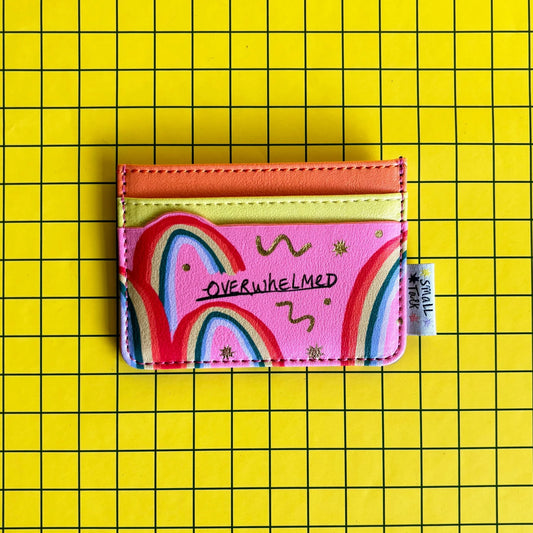 A pink, pocket-sized card holder featuring colourful rainbow designs and the word OVERWHELMED, set against a yellow grid background—ideal as a quirky gift for mates.