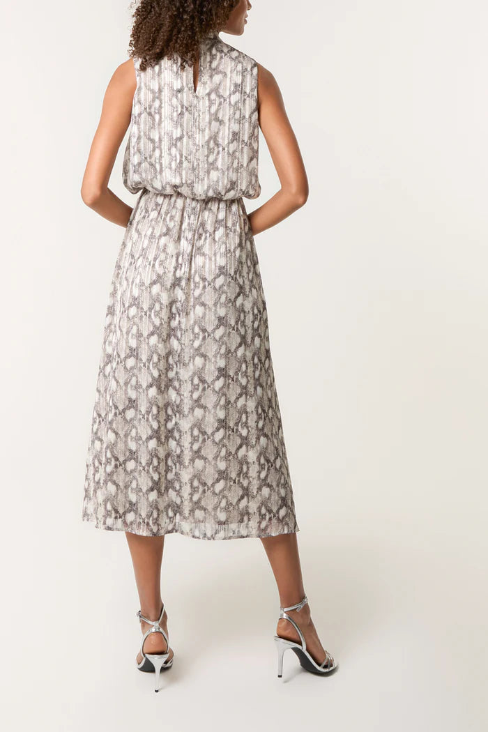 A woman stands with her back to the viewer, wearing a sleeveless, midi-length snake print dress in taupe and white, paired with silver high-heeled sandals against a plain background.