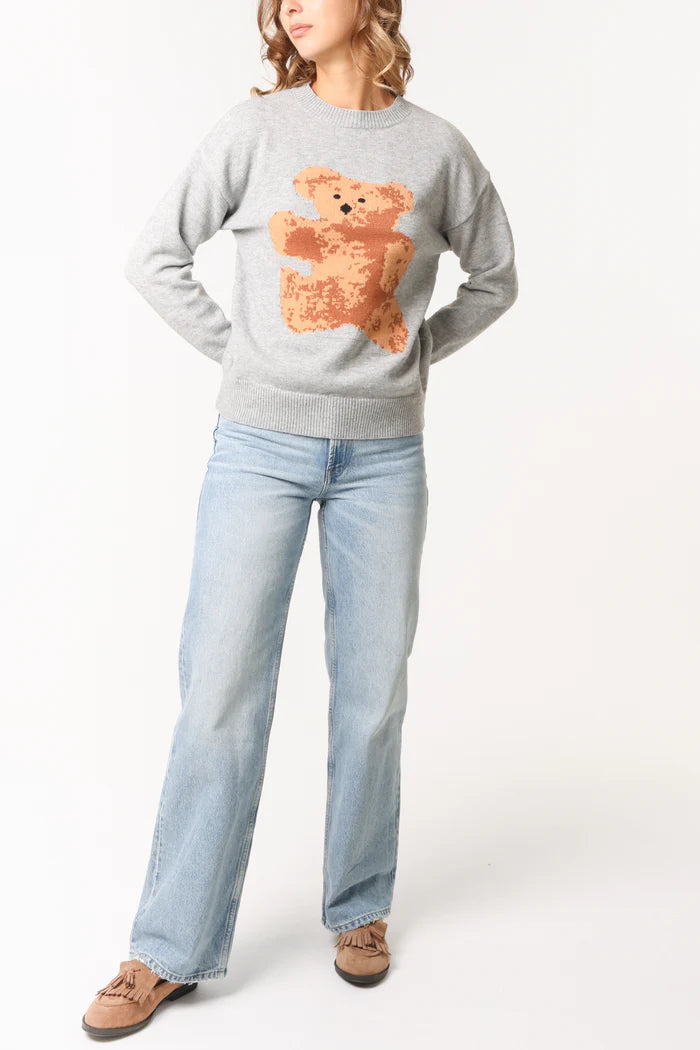 Person wearing a light grey bear jacquard jumper featuring a large brown teddy graphic, light blue jeans, and tan moccasin shoes, standing against a plain white background.