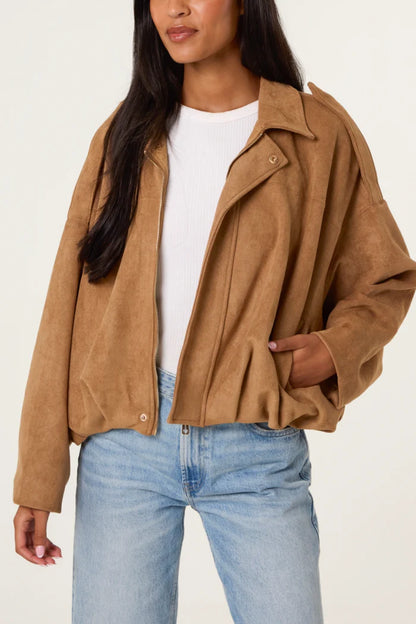 A woman dressed in a vegan-friendly tan soft-touch suedette jacket layered over a white ribbed top and light blue jeans, her hand tucked casually in her pocket.