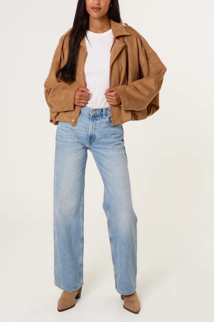 A person wearing a tan oversized soft-touch suedette jacket, white ribbed top, light blue wide-leg jeans, and tan pointed-toe boots stands against a plain background.