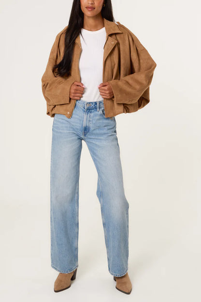 A person wearing a tan oversized soft-touch suedette jacket, white ribbed top, light blue wide-leg jeans, and tan pointed-toe boots stands against a plain background.