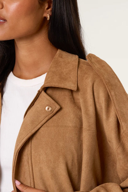 A woman in a vegan-friendly tan suede jacket over a white top, with her dark hair down, is pictured from the shoulders up against a plain background.