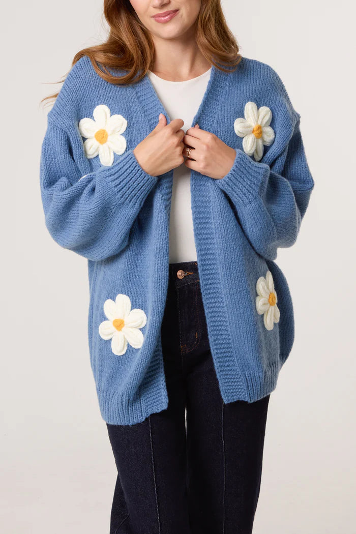 A woman wears a vegan-friendly Flocking Daisy Cardigan adorned with large white and yellow daisy appliqués, layered over a white top and dark jeans.
