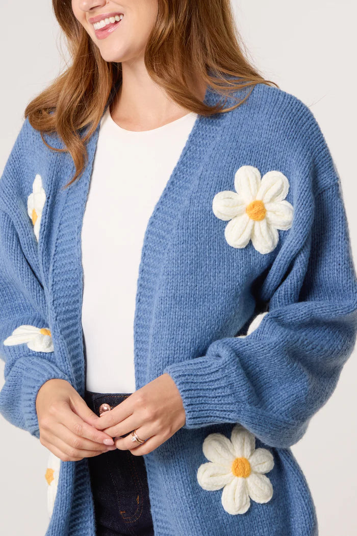 A woman wearing a vegan-friendly blue flocking daisy cardigan with large white and yellow appliqués, paired with a white top and dark trousers.