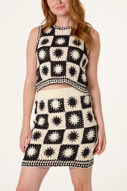 A woman wears a bohemian-inspired black and white patchwork crochet set featuring square patterns and starburst designs, standing against a plain backdrop.