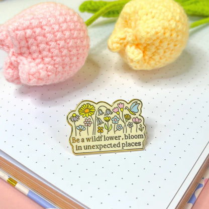 A wildflower badge bearing the phrase Be a wildflower, bloom in unexpected places rests on a dotted notebook next to two crocheted flowers.