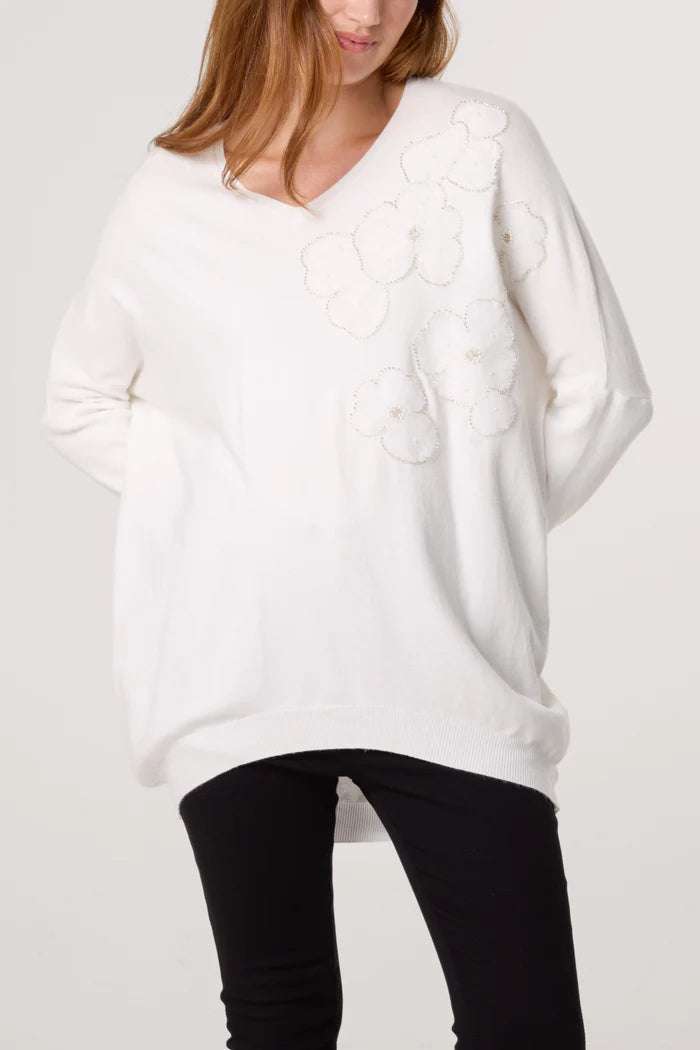 A person in a floral-embellished jumper featuring delicate embroidery on the shoulder, paired with black trousers, stands against a plain background—an effortless take on women’s fashion.