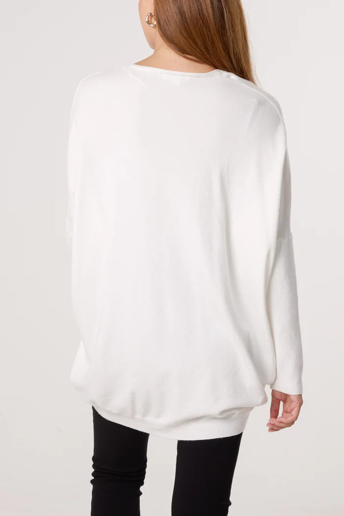 A person with long hair wears a loose-fitting white V-neck jumper and black trousers, facing away from the camera against a plain background, exemplifying timeless women's fashion.