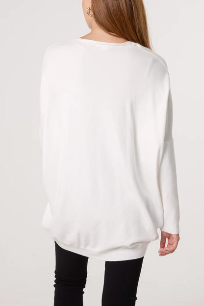 A person with long hair wears a loose-fitting white V-neck jumper and black trousers, facing away from the camera against a plain background, exemplifying timeless women's fashion.