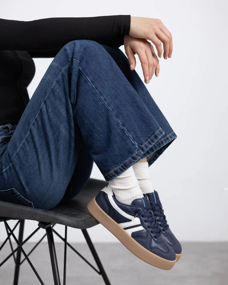 Person seated on a black chair, dressed in blue jeans, white socks, PETA-certified vegan trainers with non-slip soles and white details, and a black long-sleeved top.