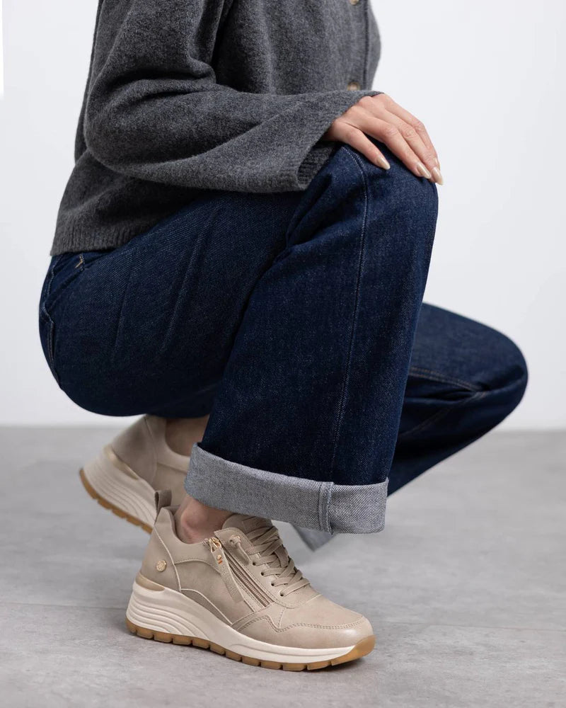 Person squatting on a grey floor, wearing rolled-up dark blue jeans, a grey jumper, and beige and gold vegan trainers with removable insoles. Only the lower half of their body is visible.