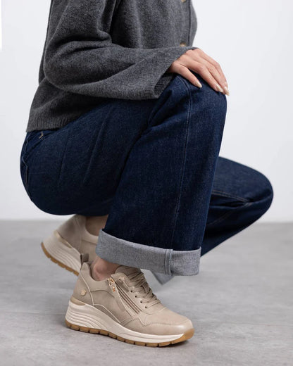 Person squatting on a grey floor, wearing rolled-up dark blue jeans, a grey jumper, and beige and gold vegan trainers with removable insoles. Only the lower half of their body is visible.