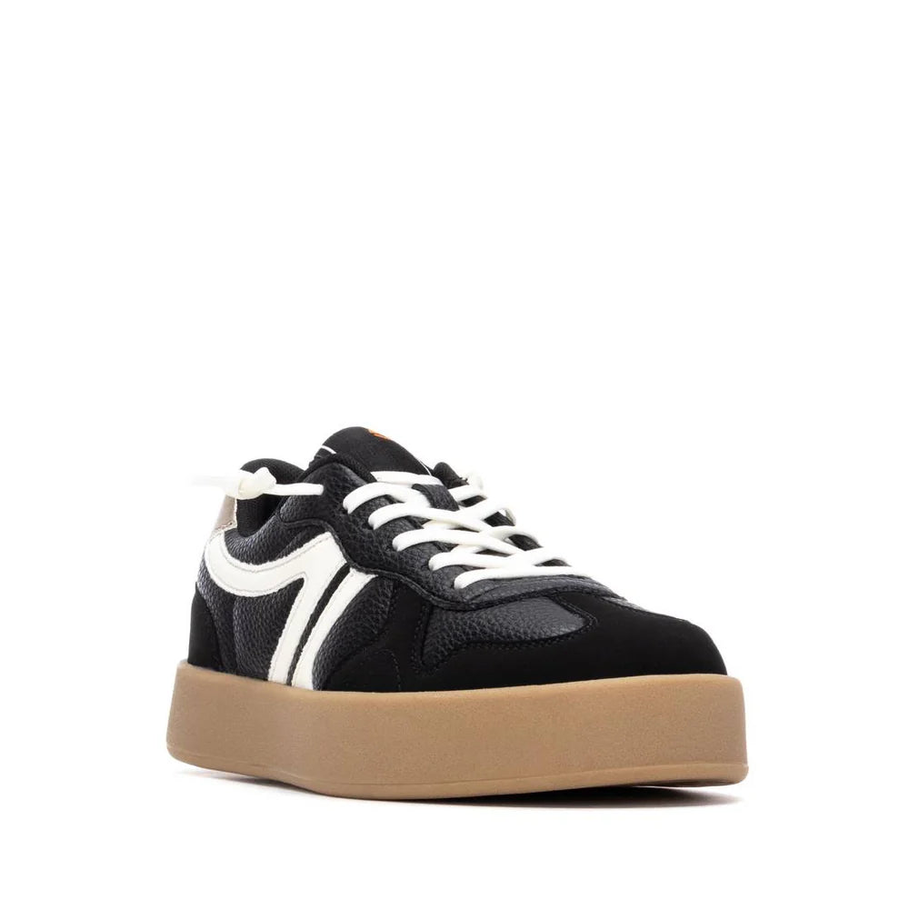A black vegan trainer with white laces and detailing, featuring a chunky tan non-slip rubber sole, photographed against a white background.